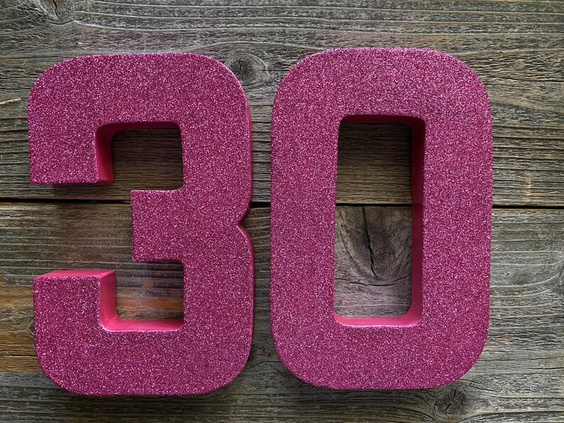 May include: Two large, pink, glitter-covered numbers, 30, are displayed on a wooden background.