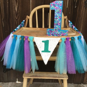 Puede incluir: Una silla alta de madera decorada con una guirnalda de tul turquesa, morado y azul. Una pancarta blanca con un n&uacute;mero verde "1" cuelga de la silla. Un gran n&uacute;mero de madera "1" con un patr&oacute;n de escamas de sirena est&aacute; en la bandeja de la silla.