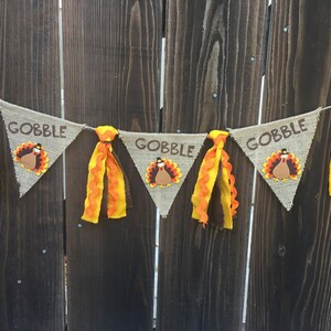 May include: Thanksgiving banner with burlap pennants. Each pennant has the word "GOBBLE" in brown letters and a turkey applique. The banner is decorated with orange and brown fabric tassels, perfect for Thanksgiving decor.