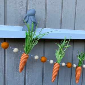 May include: A carrot garland with orange and white felt balls and green felt carrot tops. The garland is hanging from a white shelf with a gray bunny figurine on top.