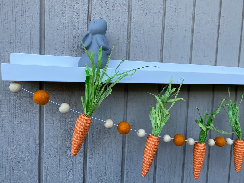 May include: A carrot garland with orange and white felt balls and green felt carrot tops. The garland is hanging from a white shelf with a gray bunny figurine on top.