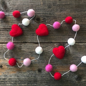 May include: A Valentine's Day garland featuring red felt hearts and colorful felt balls in shades of pink, red, and white. The garland is strung on a white string, creating a festive and decorative accent. The background is a rustic wooden surface.