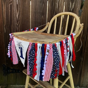 May include: A red, white, and blue fabric banner with stars and stripes hangs around a wooden high chair. The banner has a burlap flag with a firework and the number "1" on it.