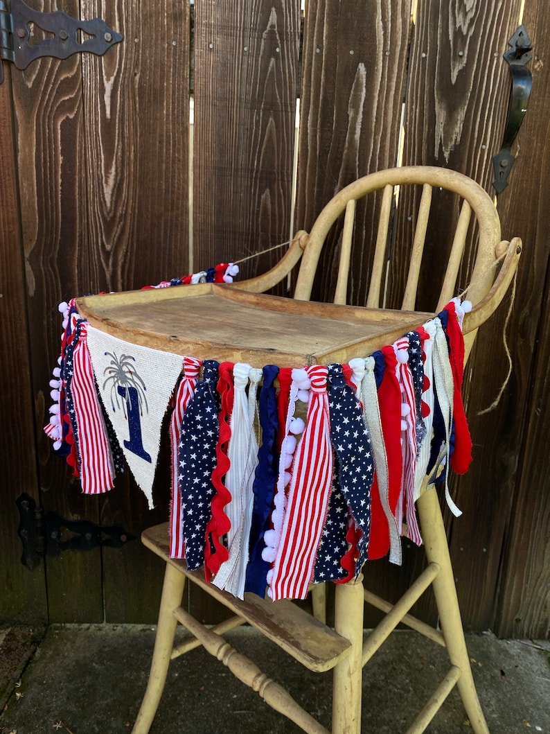 May include: A red, white, and blue fabric banner with stars and stripes hangs around a wooden high chair. The banner has a burlap flag with a firework and the number "1" on it.