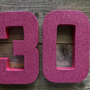 May include: Two large, pink, glitter-covered numbers, 30, are displayed on a wooden background.