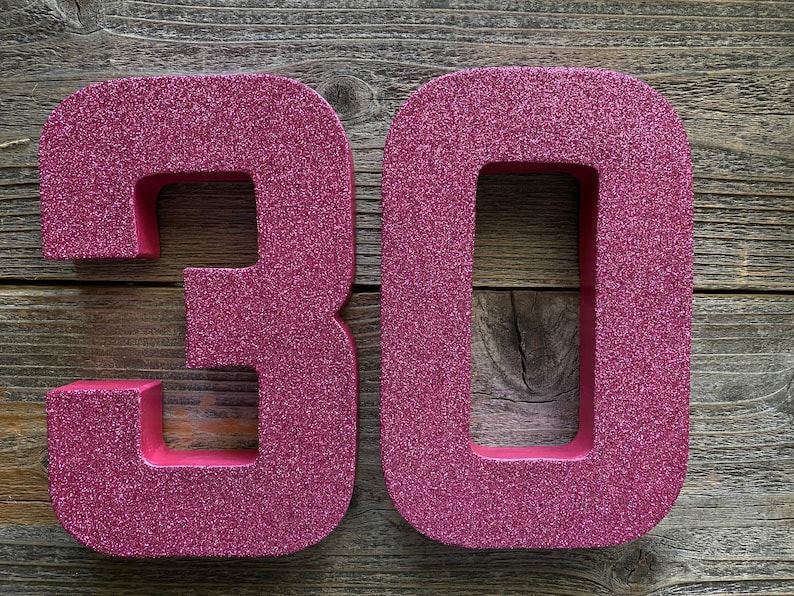 May include: Two large, pink, glitter-covered numbers, 30, are displayed on a wooden background.