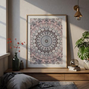 May include: A framed print featuring a detailed mandala design in shades of pink, gray, and brown. The artwork is displayed on a wooden surface, with a window and decorative items in the background. The print has a light wooden frame.