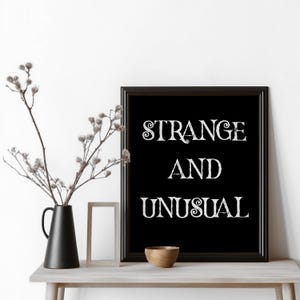 May include: A black framed print with the words "STRANGE AND UNUSUAL" in white script. The print is displayed on a light-coloured table, alongside a black vase with branches, a small wooden bowl, and a picture frame.