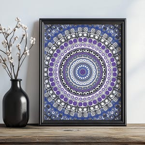 Può includere: Una stampa artistica incorniciata con un design mandala circolare nei toni del blu, viola, bianco e nero. L'opera d'arte è esposta in una cornice nera e poggia su una superficie di legno. Un vaso nero con fiori bianchi è a sinistra.