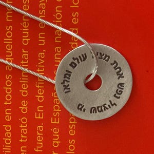 May include: A silver necklace with a round pendant inscribed with Hebrew text. The pendant is on a red surface.