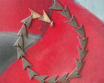 May include: A silver necklace with a geometric design. The necklace is made up of a series of triangular links that are connected together. The necklace is lying on a red and gray surface.