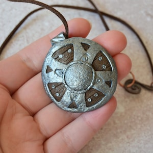 Peut inclure: Un collier pendentif rond avec un motif de bouclier argenté et des accents en bois marron. Le pendentif est attaché à un collier en cordon marron. Le bouclier a un bossage central et des rivets décoratifs.
