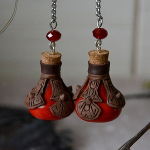 May include: A pair of red glass bottle earrings with cork stoppers and brown leather accents. The earrings are hanging from silver chains with red gemstone beads.