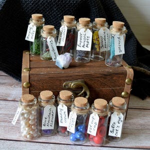 Könnte beinhalten: Eine Sammlung kleiner Glasflaschen mit Korken, die verschiedene Gegenstände wie Perlen, Moos und bunte Perlen enthalten. Die Flaschen sind mit handgeschriebenen Etiketten versehen und auf einer Holzkiste angeordnet.