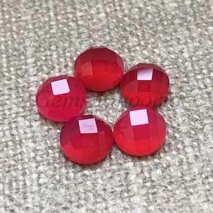 May include: Six round, red gemstone cabochons with faceted edges. The cabochons are arranged in a circle on a textured, light brown surface.