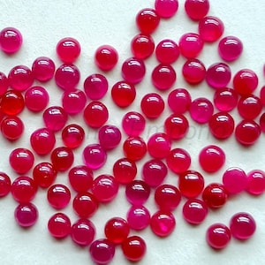 Peut inclure: Une collection de pierres précieuses rondes en cabochon rose. Les cabochons sont éparpillés sur une surface blanche.