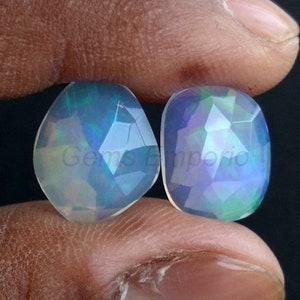 Op de afbeelding: Twee regenboogkleurige opaal edelstenen, geslepen in een gefacetteerde, druppelvorm. De stenen zijn van een lichtblauwe kleur met groene en blauwe schittering.