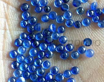 May include: A collection of small, round, deep blue gemstones scattered across a light-colored surface. The gems have a polished, reflective appearance, with a slight variation in color and size. The overall effect is a display of vibrant, sparkling blue stones.