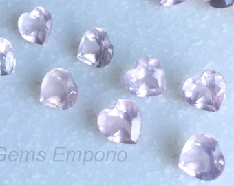 May include: A collection of pale pink heart-shaped gemstones. The gemstones are scattered on a light blue surface. Gems Emporio is written in black text at the bottom of the image.