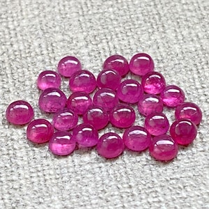 May include: A group of 25 round, pink ruby gemstones. The gemstones are smooth and polished, and they are arranged in a loose pile.