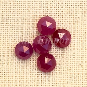 May include: Six faceted ruby red gemstones, each with a pointed top, arranged in a cluster on a beige fabric background.