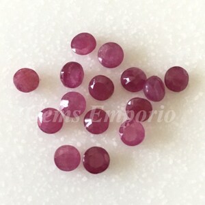 Könnte beinhalten: Eine Ansammlung facettierter, runder, tiefroter Edelsteine. Die Edelsteine sind auf einer weißen Oberfläche verstreut. Der Text "Gems Emporio" ist teilweise sichtbar. Diese Edelsteine könnten für die Schmuckherstellung verwendet werden.