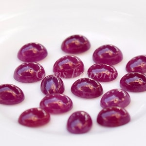 May include: A collection of 16 round, cabochon cut, ruby red gemstones. The gemstones are arranged on a white surface.