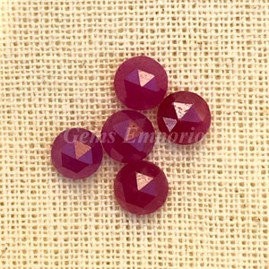Könnte beinhalten: Fünf facettierte rubinrote Edelsteine mit goldfarbener Oberfläche. Die Edelsteine sind rund und haben ein facettierendes Design. Die Edelsteine befinden sich auf einem beigefarbenen Stoffhintergrund.