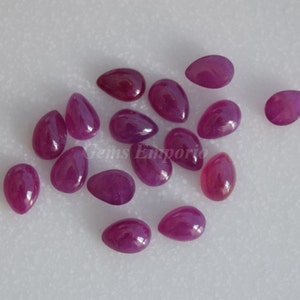 May include: A collection of 14 pear-shaped ruby gemstones, ranging in size and shade of pink. The stones are arranged on a white surface.