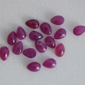 May include: A group of 14 pear-shaped ruby gemstones, each with a deep pink hue and a glossy finish. The stones are scattered on a white surface.