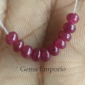 Op de afbeelding: Een streng van acht ronde, rode edelsteen kralen geregen aan een doorzichtig elastisch koord. De kralen zijn ongeveer 4 mm in diameter. De kralen zijn in een halve cirkelvorm gerangschikt. De tekst "Gems Emporio" is zichtbaar onder de kralen.