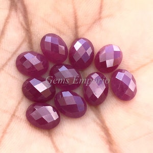 May include: A collection of ten faceted oval ruby gemstones in various shades of pink and red. The gemstones are displayed on a hand.