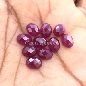 May include: Ten faceted oval ruby gemstones in various shades of pink and red. The gemstones are arranged in a loose pile on a hand.
