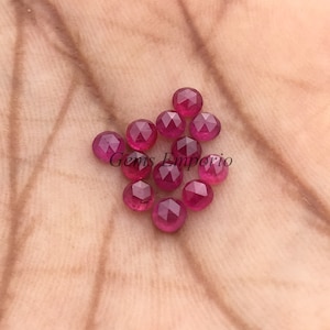 May include: A collection of faceted, round, deep red gemstones. The gems are scattered on a light brown surface, with the text "Gems Emporio" visible. The gemstones have a polished, reflective surface, and are likely rubies.