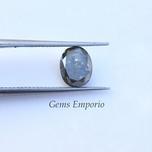 Puede incluir: Un diamante gris oscuro de forma ovalada sostenido por unas pinzas. El diamante tiene un corte facetado y est&aacute; colocado sobre un fondo blanco. El texto "Gems Emporio" es visible debajo del diamante.