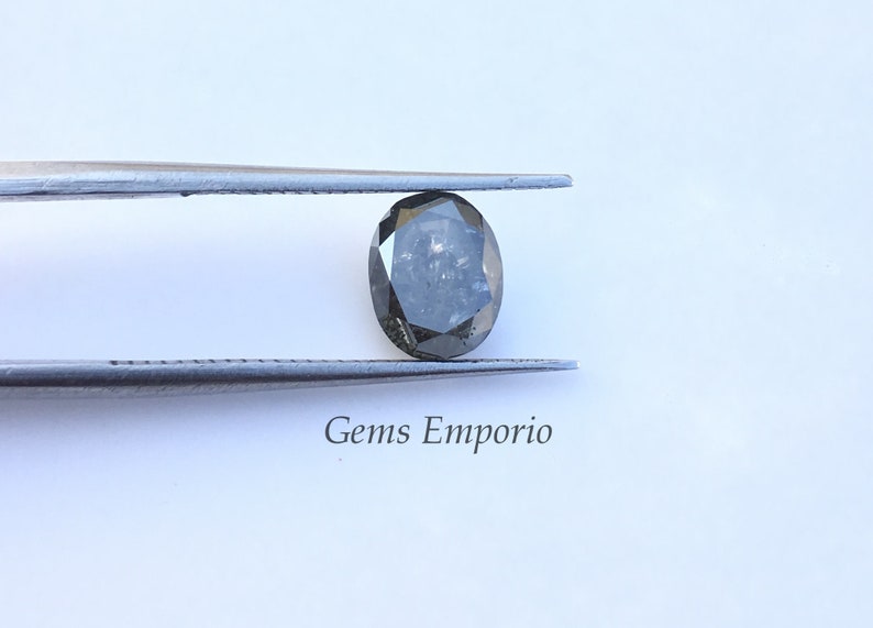 Puede incluir: Un diamante gris oscuro de forma ovalada sostenido por unas pinzas. El diamante tiene un corte facetado y est&aacute; colocado sobre un fondo blanco. El texto "Gems Emporio" es visible debajo del diamante.