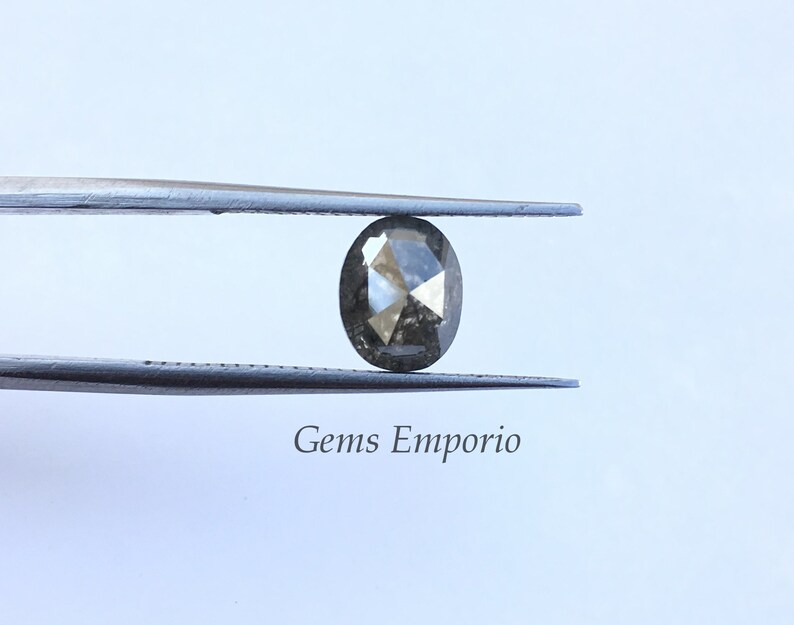 Puede incluir: Un diamante gris oscuro de forma ovalada con un corte facetado, sostenido por unas pinzas. Gems Emporio.