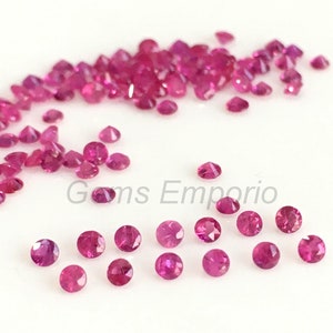 May include: A scattering of small, round, pink ruby gemstones. The gemstones are faceted and have a brilliant shine.  Gems Emporio is printed on the white surface.