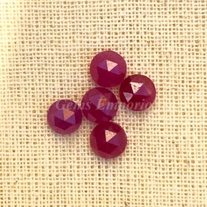 May include: Five faceted ruby red gemstones with a gold-toned finish. The gemstones are round and have a faceted design. The gemstones are on a beige fabric background.