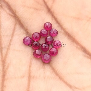 Pode incluir: Uma coleção de pedras preciosas facetadas, redondas, de cor vermelha escura. As gemas estão espalhadas sobre uma superfície castanha clara, com o texto "Gems Emporio" visível. As pedras preciosas têm uma superfície polida e refletora, e são provavelmente rubis.