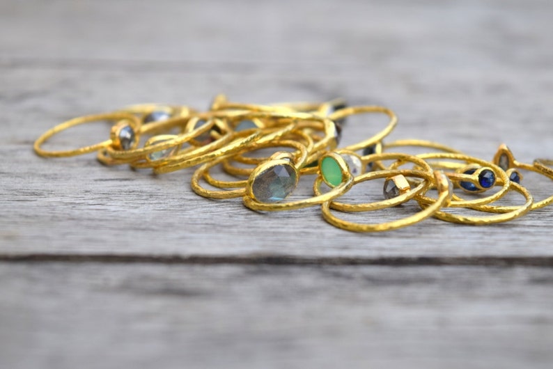 Pu&ograve; includere: Una collezione di anelli d'oro con varie pietre preziose colorate, tra cui verde, blu e grigio. Gli anelli sono impilati uno sopra l'altro su una superficie di legno.