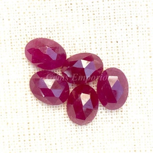 May include: Five faceted oval ruby gemstones in various shades of red. The gemstones are arranged in a cluster on a white fabric background.