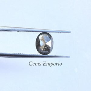 Puede incluir: Un diamante gris de forma ovalada con un corte facetado, sostenido por unas pinzas. El diamante est&aacute; colocado sobre un fondo blanco.