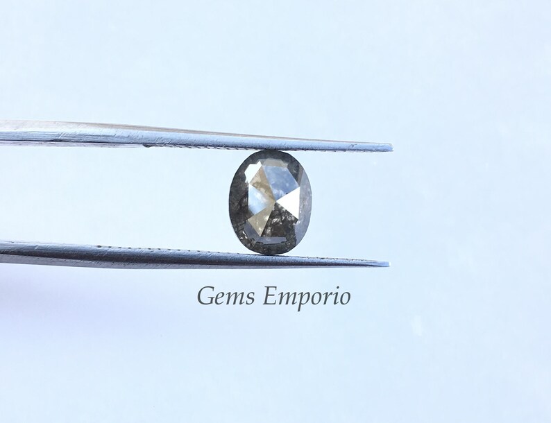 Puede incluir: Un diamante gris de forma ovalada con un corte facetado, sostenido por unas pinzas. El diamante est&aacute; colocado sobre un fondo blanco.