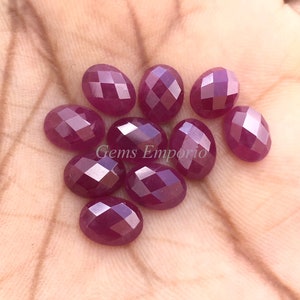 May include: A collection of oval-shaped, faceted ruby gemstones. The gems are a deep red color and have a polished, reflective surface. The image is taken from a close-up perspective, highlighting the details of the gemstones. The text "Gems Emporio" is visible.
