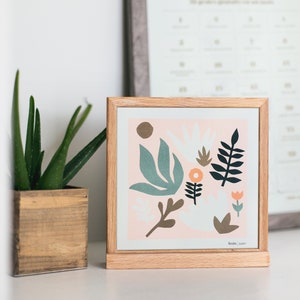 Puede incluir: Una impresi&oacute;n enmarcada con un fondo rosa y un dise&ntilde;o floral abstracto colorido. La impresi&oacute;n presenta hojas y flores verdes, azules, naranjas y marrones. El marco es de madera clara.