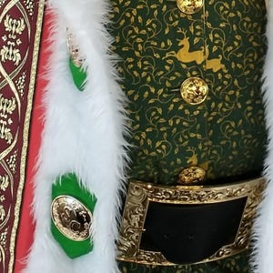 Puede incluir: Primer plano de un disfraz festivo verde y dorado. La chaqueta presenta patrones dorados de ciervos y hojas, botones dorados y una hebilla de cinturón dorada decorativa. El ribete de piel sintética blanca y un emblema verde y dorado se suman al tema festivo.