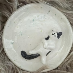 May include: A white ceramic dish with a sleeping cat figurine. The cat has black markings on its head and tail. The dish has a speckled design with green and blue accents.