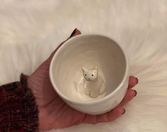 May include: A white ceramic bowl with a small, white cat figurine inside. The bowl is held in a hand against a fluffy, white background. The cat has black eyes and is sitting upright.