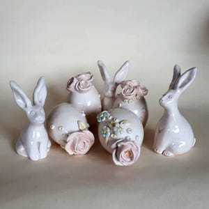 Op de afbeelding: Een verzameling keramische paashaasfiguren in verschillende poses. Sommige konijnen zijn effen, andere versierd met roze en blauwe bloemaccenten. De figuren hebben een zachte, pastelroze kleur.